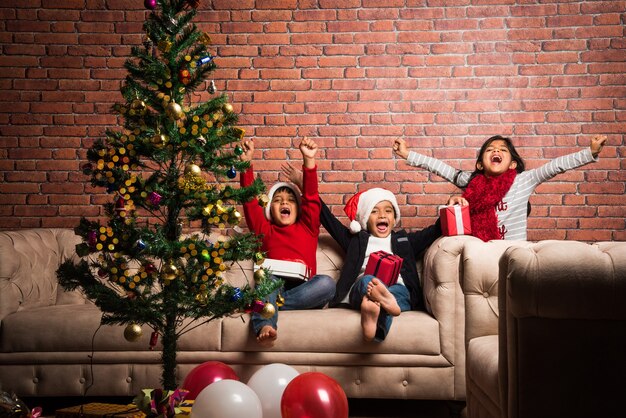 Süße kleine indische asiatische Kinder feiern Weihnachten zu Hause mit Weihnachtsmütze, Geschenken und Weihnachtsbaum