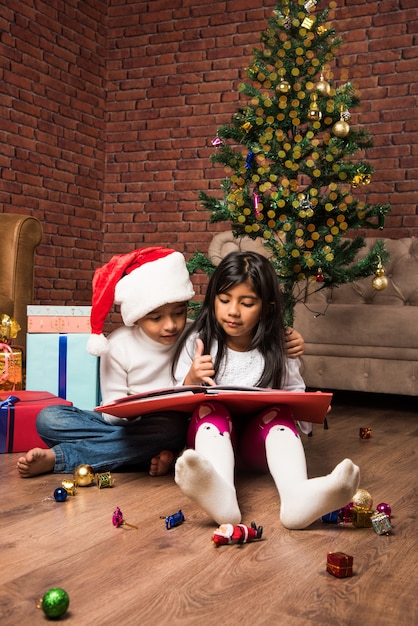 Süße kleine indische asiatische Kinder feiern Weihnachten zu Hause mit Weihnachtsmütze, Geschenken und Weihnachtsbaum