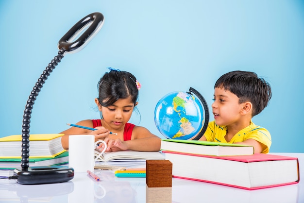 Süße kleine indische oder asiatische Kinder, die auf einem Studientisch mit Bücherstapel, Bildungsglobus, isoliert über hellblauer Farbe