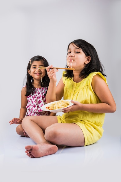 Süße kleine indische oder asiatische verspielte Mädchen, die leckere chinesische Nudeln mit Gabel oder Stäbchen essen, einzeln auf buntem Hintergrund