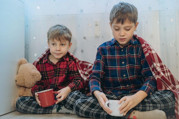 Süße kleine Jungs in Plaid gehüllt, die zu Hause mit Teddybär und kalten Tassen an der Heizung sitzen