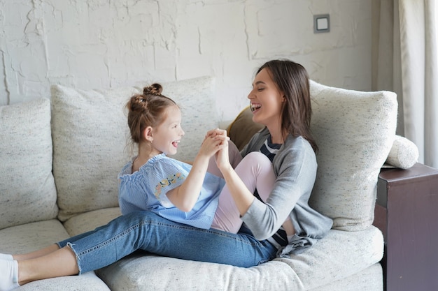 Süße kleine Tochter, die mit Mutter auf dem Sofa lacht, glückliche Mutter, die sich entspannt, Spaß mit lustigen kleinen Mädchen hat, die gemeinsam Freizeit im Wohnzimmer genießen.