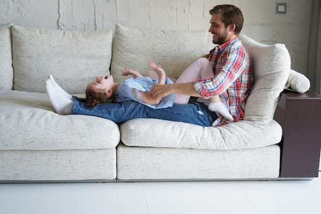 Süße kleine Tochter lacht Kitzeln spielen mit Papa auf dem Sofa, glücklicher Vater entspannt Spaß mit lustigen kleinen Kindermädchen, die gemeinsam Freizeit im Wohnzimmer genießen.