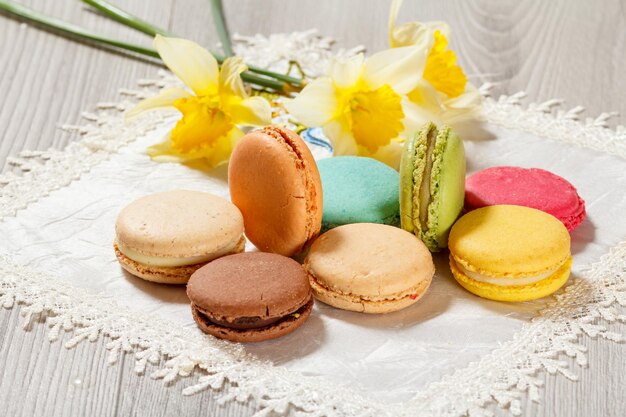 Süße Macarons Kuchen unterschiedlicher Farbe auf Seidenserviette mit Strauß frischer gelber Narzissen auf Holzhintergrund.