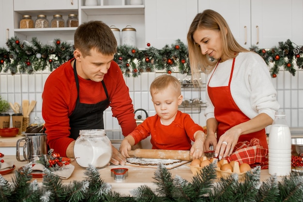 Süße schöne Familie, die Weihnachtskekse in der Küche kocht Ein kleiner Junge im roten Pullover rollt den Teig für Lebkuchen des neuen Jahres aus