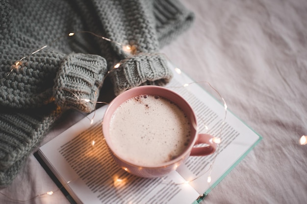 Süße Tasse Kaffee mit Weihnachtsbeleuchtung auf offenem Buch mit warmem Strickpullover im Bett aus nächster Nähe. Guten Morgen. Frühstück. Winterferienzeit.