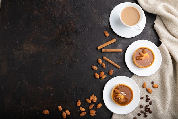 Süße Törtchen mit Mandeln und Karamellcreme mit Tasse Kaffee auf schwarzem Betonhintergrund und Leinentextil. Draufsicht, flache Lage, Kopierraum.