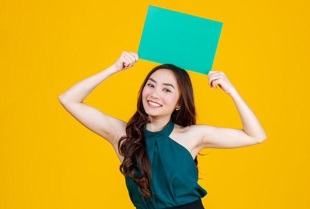 Süße und hübsche lockige Haare Asiatische weibliche Brünette, die grünes leeres Brett hält, posiert mit einer fröhlichen und positiven Geste für Werbezwecke zur Kamera, Studioaufnahme einzeln auf gelbem Hintergrund.