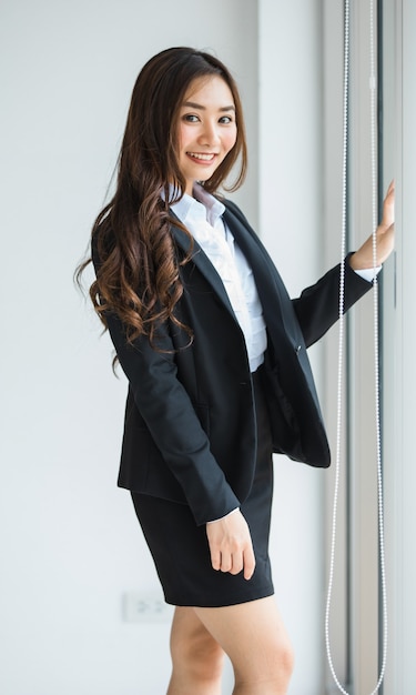 Süße und schöne brünette asiatische Unternehmerin, die wunderschön in schwarzem, edlem Anzug aussah, der im Büro steht und mit freundlichem und positivem, glücklichem Gesicht in die Kamera schaut.