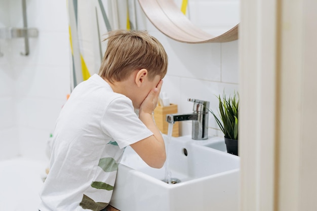 süßer 6-jähriger Junge, der sein Gesicht über dem Waschbecken im Badezimmer wäscht Bild mit selektivem Fokus