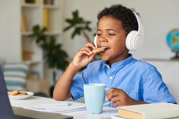 Süßer afroamerikanischer Junge mit drahtlosen Kopfhörern, der am Schreibtisch sitzt und Kekse isst, während er edu beobachtet