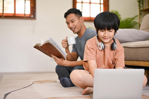 Süßer asiatischer kleiner Junge, der einen Laptop benutzt, während sein Vater hinten ein Buch liest