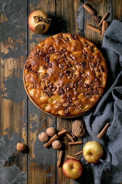 Süßer hausgemachter Apfel- und Walnusskuchen Charlotte auf Teller mit frischen Gartenäpfeln, Zimt und Nüssen auf dunklem Holzbretthintergrund. Herbstliche Hausmannskost. Flach legen, Platz kopieren