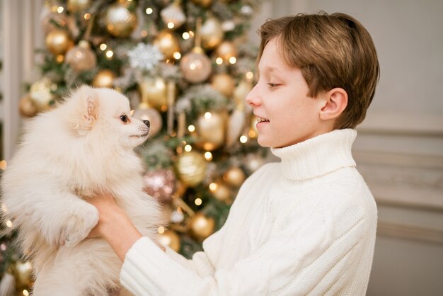 Süßer Junge am Weihnachtsbaum mit Hund in Erwartung der frohen Weihnachten, glücklicher Junge und Hund umarmt ihr T...