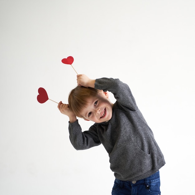 Süßer Junge, der mit zwei roten Papierherzen auf Stöcken spielt, scherzt medizinisches Konzept des Gesundheitswesens
