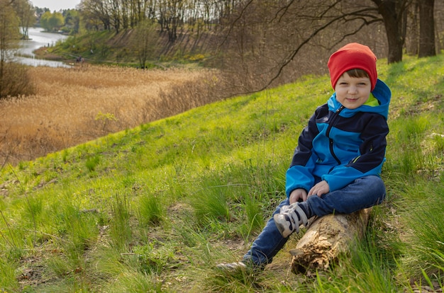 Süßer Junge Ein vierjähriger Junge sitzt auf einem Hügel auf einem Baumstamm und schaut auf den See. Grüner Familientourismus, Wandern mit Kindern im Freien