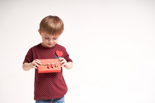 Süßer Junge hält rotes Geschenk mit Herzen auf weißem Hintergrund Valentinstag oder Kindergesundheitsmedi...