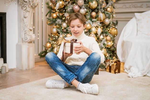 Süßer Junge im Pullover sitzt am Weihnachtsbaum mit Geschenken Kaukasischer Teenager zu Hause mit festlichem Ne...