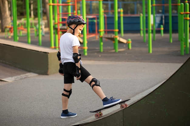 Süßer Junge in einem Helm, der in einem speziellen Bereich im Skatepark steht und auf Skateboard tritt Kind führt Tricks durch Sommersport-Aktivitätskonzept Glückliche Kindheit