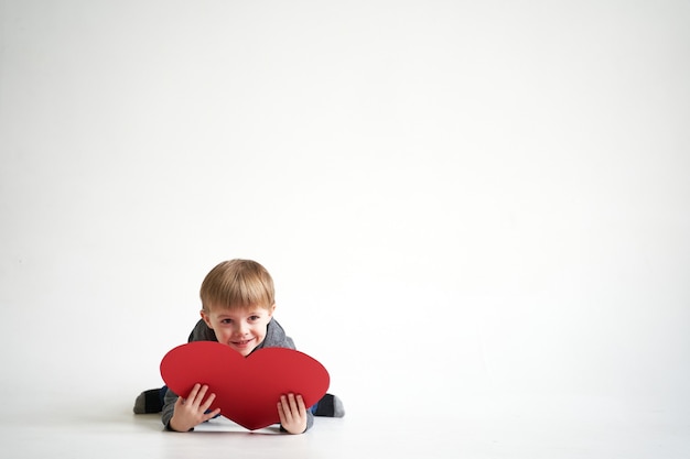 Süßer Junge liegt mit rotem Papierherz auf weißem Hintergrund Valentinstag oder Kindergesundheitsmedizin...