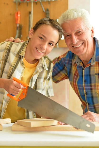 Süßer Junge mit Großvater, der zu Hause mit Holzbohlen und Handsäge arbeitet