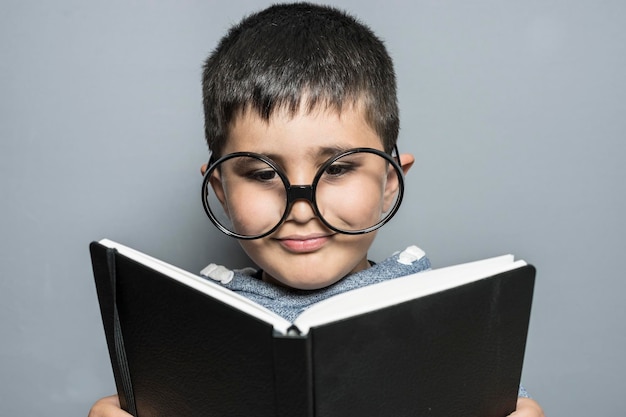 Süßer Junge mit riesiger Brille, der ein Buch mit lustigen und abwechslungsreichen Gesten liest