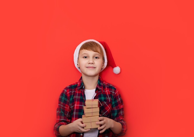 Süßer kaukasischer Teenager-Junge in karierten Shorts hält Geschenkboxen auf rotem Hintergrund Platz für Design oder Text kopieren Banner Weihnachten Neujahr Mockup-Vorlage