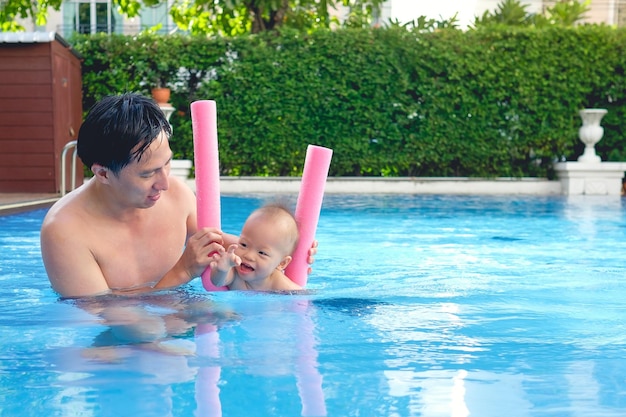 Foto süßer kleiner asiate, 18 monate, 1 jahr alt, kleinkind, junge, kind, das lernt, mit poolnudeln mit papa im außenpool zu schwimmen