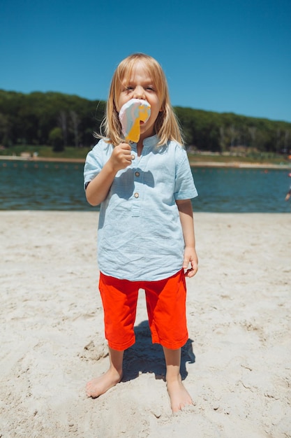 Süßer kleiner blonder Junge mit langen Haaren, der im Strandsommerurlaub Eis isst
