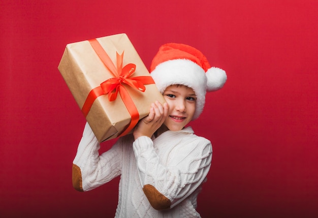 Süßer kleiner Junge in einem Weihnachtsmann-Hut, der eine Geschenkbox auf rotem Hintergrund hält, ein Kind mit einem Neujahrsgeschenk für Weihnachten, Neujahr