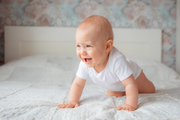 Süßer kleiner Junge in einem weißen Body, der im Schlafzimmer auf dem Bett krabbelt, das Konzept der glücklichen Mutterschaft