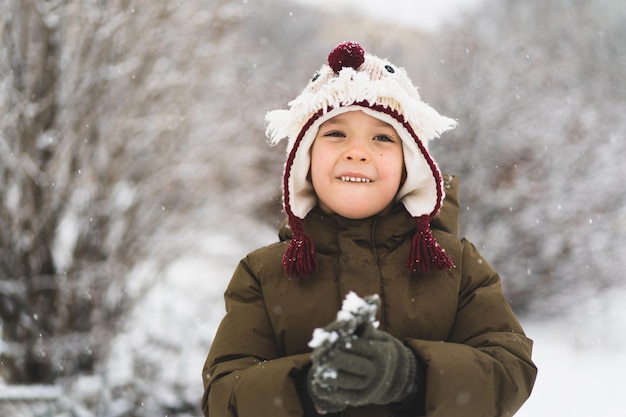 Süßer kleiner Junge mit lustigem Winterhut geht während eines Schneefalls im Freien Winteraktivitäten für Kinder