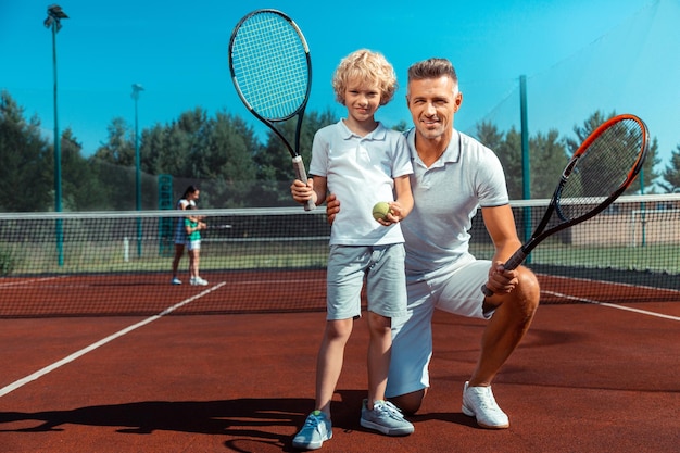 Süßer, lockiger Sohn, der sich glücklich fühlt, während er mit Papa Tennis spielt