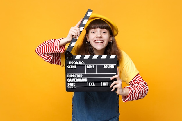 Süßer Mädchen-Teenager in französischer Baskenmütze, Denim-Sommerkleid mit klassischer schwarzer Filmklappe isoliert auf gelbem Wandhintergrund. Menschen aufrichtige Emotionen Lifestyle-Konzept. Mock-up-Kopienbereich.