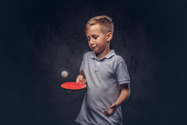 Süßer rothaariger Junge in einem weißen T-Shirt, der in einem Studio Tischtennis spielt. Getrennt auf einem dunklen strukturierten Hintergrund.