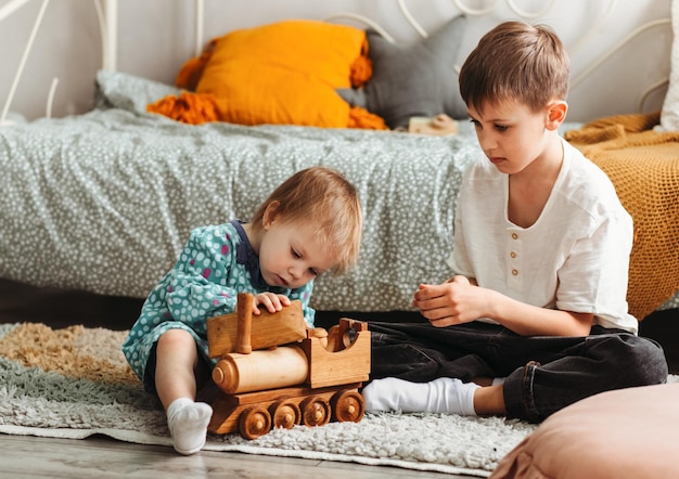 Süßer Teenager und seine kleine Schwester sitzen auf einem flauschigen Teppich auf dem Boden des Wohnzimmers und spielen nach dem Abendessen mit Holzeisenbahn