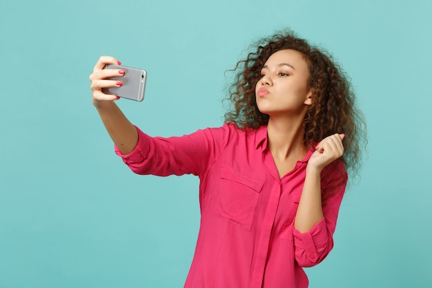 Süßes afrikanisches Mädchen in Freizeitkleidung bläst Luftkuss, macht Selfie-Schuss auf dem Handy einzeln auf blauem türkisfarbenem Hintergrund. Menschen aufrichtige Emotionen, Lifestyle-Konzept. Kopieren Sie Platz.