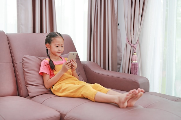 Süßes asiatisches kleines Mädchen, das zu Hause Smartphone auf dem Sofa im Wohnzimmer spielt