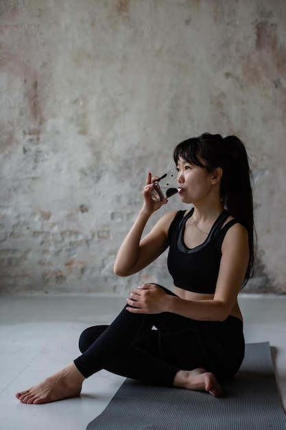 Süßes asiatisches Mädchen mit dunklen langen Haaren in Trainingssportbekleidung, das sich nach einem Trainingstrinkwasser ausruht