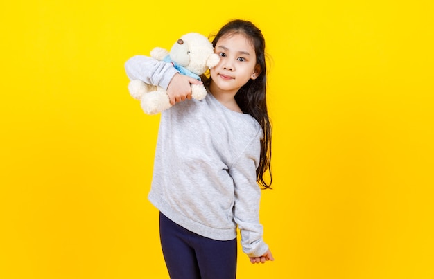 Süßes asiatisches Mädchen mit Pullover, Porträtfoto, das schöne Bärenpuppe auf gelbem Hintergrund umarmt.