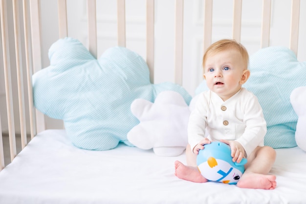 Süßes Baby 6 Monate alter blonder Junge in einer Krippe in einem hellen Schlafzimmer in einem weißen Baumwollbody mit einem blauen Spielzeug das Konzept der Kinderartikel