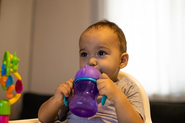 Süßes Baby, das im Hochstuhl sitzt und Wasser aus der Babyflasche trinkt Baby zu Hause