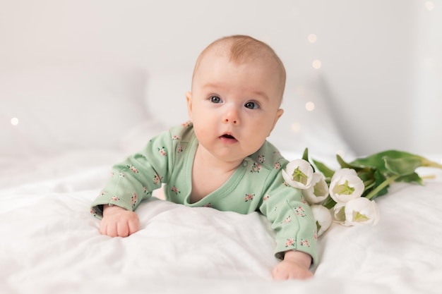 Foto süßes baby in einem grünen baumwollbody zu hause auf einem weißen bett mit tulpen. frühling, muttertag, märz