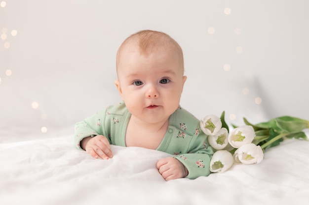 Süßes Baby in einem grünen Baumwollbody zu Hause auf einem weißen Bett mit Tulpen. Frühling, Muttertag, März