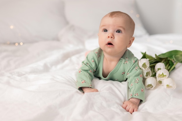 Süßes Baby in einem grünen Baumwollbody zu Hause auf einem weißen Bett mit Tulpen. Frühling, Muttertag, März