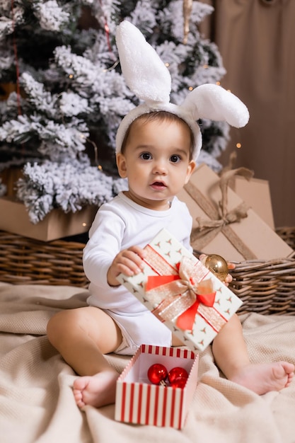 Süßes Baby in einem weißen Bodysuit und Hasenohren sitzt neben dem Weihnachtsbaum zu Hause und betrachtet Geschenke Symbol des Jahres Das Jahr des Kaninchens 2023