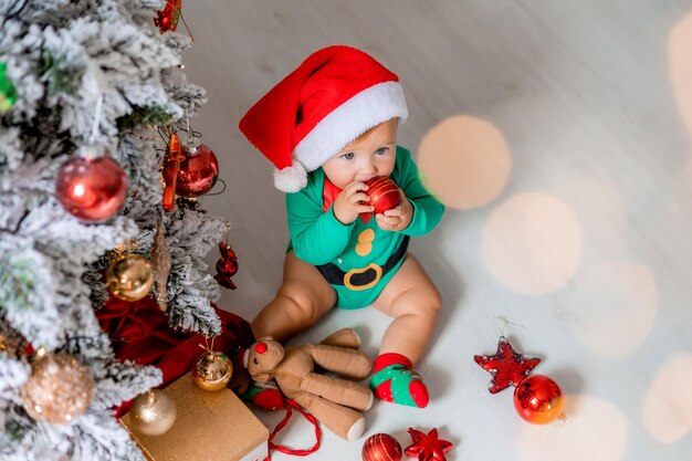 Süßes Baby in grünem Strampler und Weihnachtsmütze schmückt den Weihnachtsbaum mit bunten Luftballons