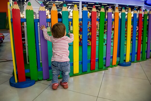 Süßes Baby steht am bunten Zaun des Kinderspielzimmers und möchte mit dem Finger nach innen spielen