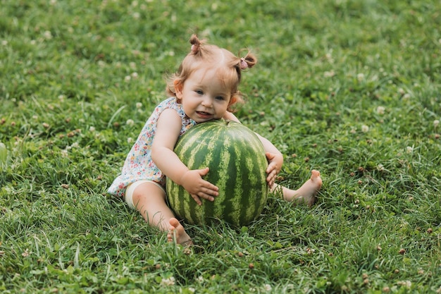 Süßes Babymädchen mit einer riesigen Wassermelone sitzt auf einem grünen Rasen. Kind spielt im Freien