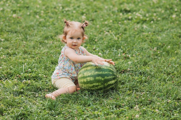 Süßes Babymädchen mit einer riesigen Wassermelone sitzt auf einem grünen Rasen. Kind spielt im Freien